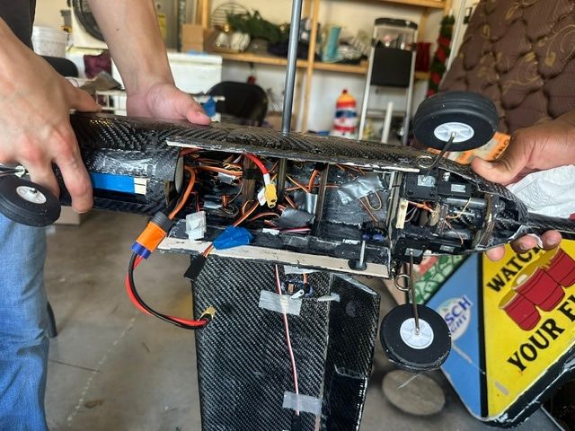 The fuselage held open by two people, showing servos, wiring, a battery and connectors packed inside the carbon shell, with the wheels attached.