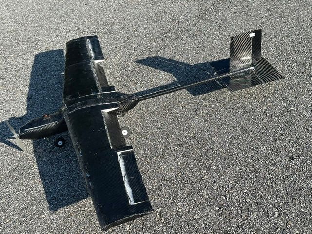 The carbon-fibre UAV resting on asphalt in low sun, seen from above and behind the left wing. A single carbon tube runs from the wing to the tail surfaces; a two-blade propeller sits at the nose.